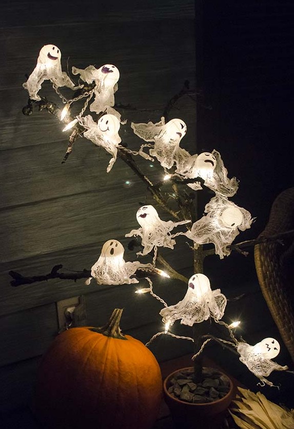 Halloweenske dekorácie zo svetelnej reťaze — svietiaci duchovia z bielej gázy na konári nad tekvicou