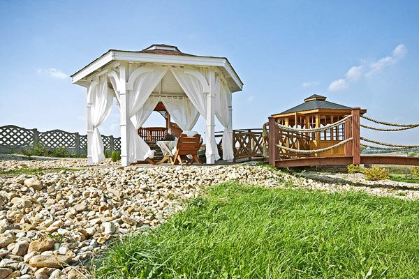 Drevený altánok FLORIDA – biely so závesmi na kamenistom svahu
